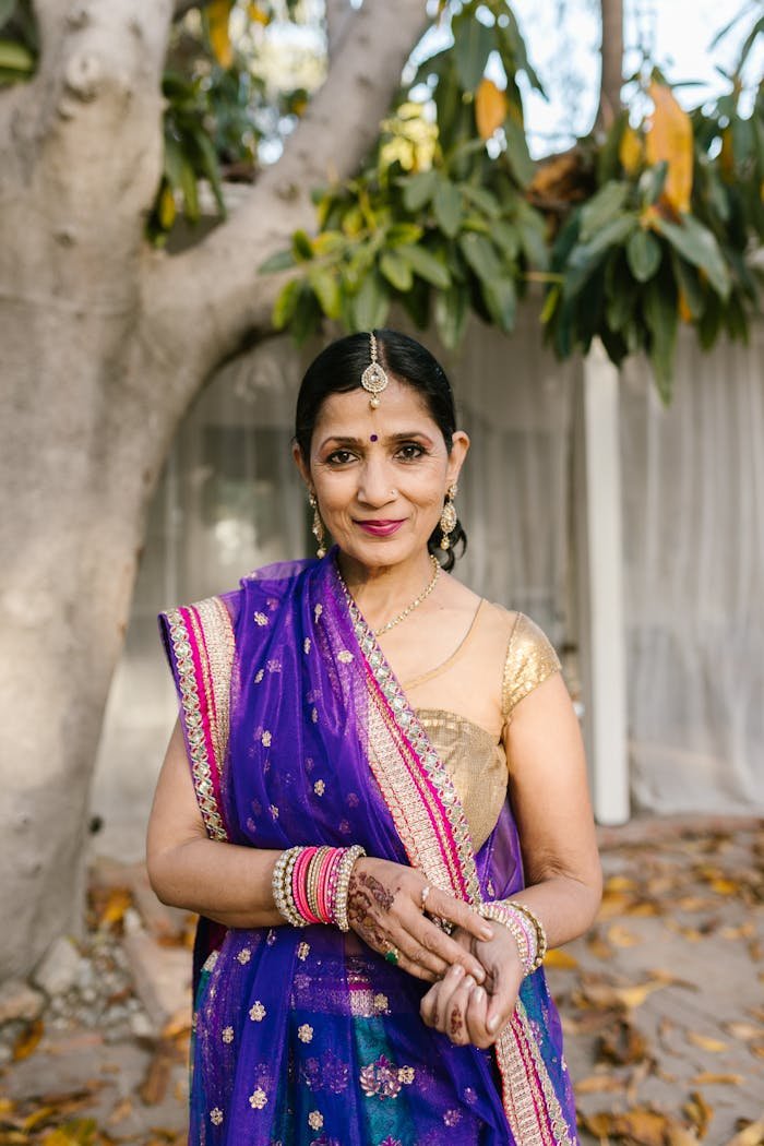 Graceful Indian woman in a vibrant saree and jewelry, standing outdoors with trees.